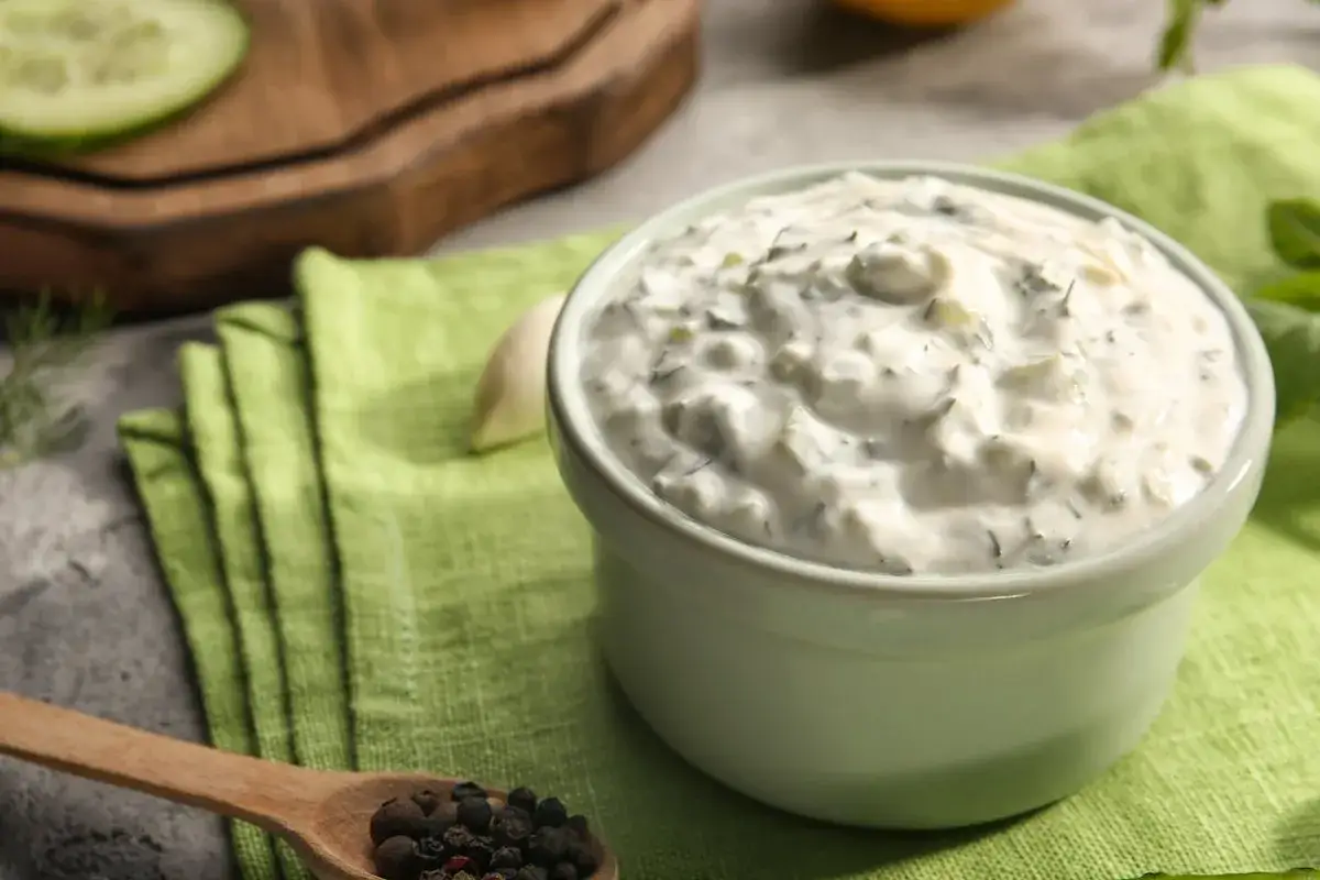 Una ciotola di cremosa salsa yogurt con erbe fresche, accanto a pepe nero e aglio. Perfetta per accompagnare ogni piatto.