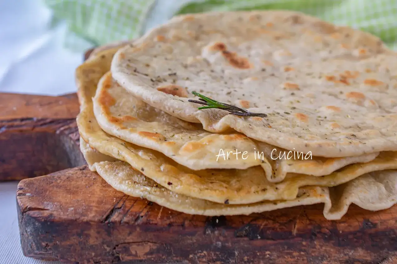 Pila di piadine senza strutto, condite con erbe aromatiche, su un tagliere di legno.