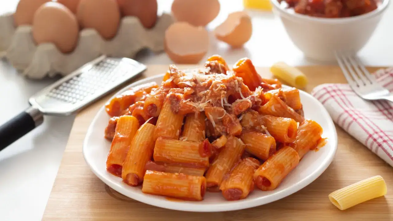 Un piatto di pasta alla zozzona fumante, con sugo ricco e formaggio grattugiato. Ingredienti freschi come uova e una grattugia completano la scena.