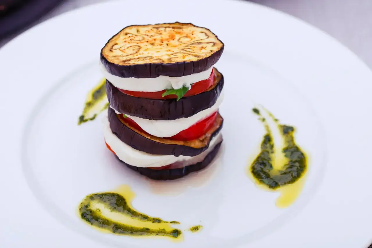 Parmigiana di melanzane grigliate a strati con mozzarella e pomodoro, guarnita con pesto su piatto bianco.