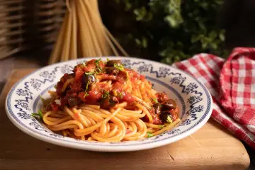 Piatto di spaghetti alla puttanesca, conditi con pomodoro, olive e capperi, guarniti con prezzemolo fresco.