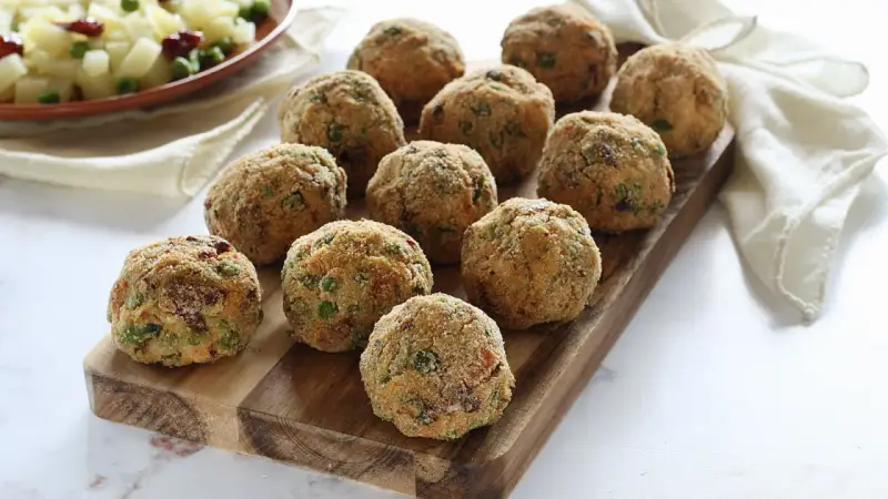 Deliziose polpette di verdure dorate e croccanti, disposte su un tagliere di legno. Un contorno di patate e piselli sullo sfondo.