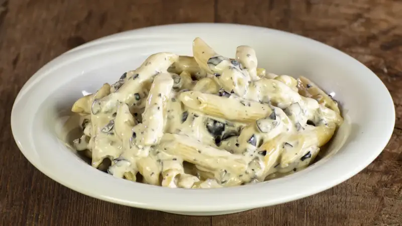 Penne cremose con pezzetti scuri, un piatto invitante di pasta alla cenere, servito in una ciotola bianca su un tavolo di legno rustico.